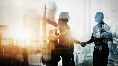 Zwei silhouettes Geschäftspersonen schütteln sich die Hände vor einer industriellen Kulisse mit Gebäuden und Kraftwerken; die Szene symbolisiert Zusammenarbeit und eine geschäftliche Vereinbarung.