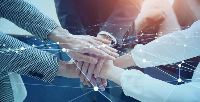 Mehrere Hände verschiedener Personen legen sich übereinander zu einem Teamzeichen; Symbol für Zusammenarbeit, Zusammenhalt und gemeinsames Engagement, vor einem Hintergrund abstrakter Netzlinien, die Vernetzung und Kommunikation darstellen.
