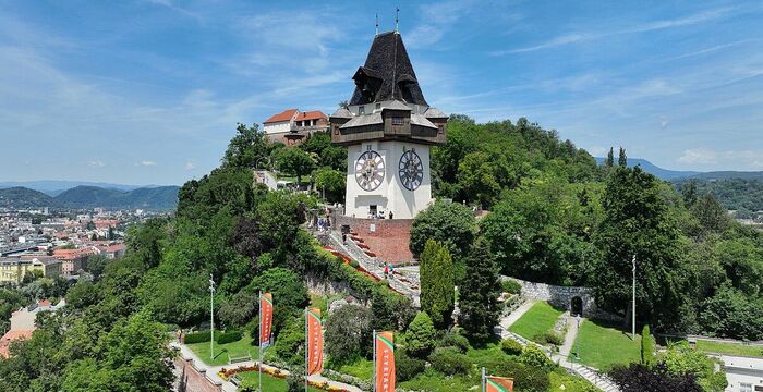 Auf einem bewaldeten Hügel steht ein historischer Uhrturm mit zwei Zifferblättern in einem weißen Aufbau und dunklem Turmhelm. Rund um den Turm eine gepflegte Parkanlage mit orangen Fahnen. Im Hintergrund eine Stadtlandschaft und sanfte Hügel unter blauem Himmel.