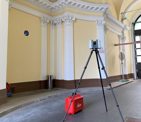 Historischer Innenraum mit gelben Wänden und weißen Säulen. Vor einem Stativ mit Messgerät steht eine rote Transportbox; im Hintergrund rechts ist eine Absperrung und ein Fenster sichtbar.