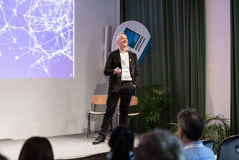 Ein Mann in business-ähnlicher Kleidung hält einen Vortrag auf einer Bühne vor einem Publikum. Links ist eine Projektion mit einem abstrakten Netzwerk-Muster zu sehen; im Hintergrund stehen Vorhänge, zwei Stühle, ein Banner und Pflanzen.