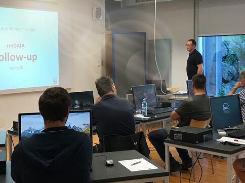 Eine Schulungs- oder Seminarraum-Szene: Ein Vortragender steht vorne neben einem Laptop und präsentiert eine Folie; mehrere Teilnehmer sitzen mit Laptops an Tischen, schauen zum Bildschirm. Fenster im Hintergrund, Wasserflasche auf dem Tisch.