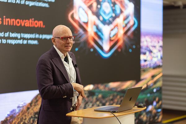 Ein Mann in dunklem Anzug und Brille steht hinter einem runden Stehpult mit Laptop und spricht in ein Mikrofon. Im Hintergrund zeigt eine große Präsentationswand digitale Grafiken und das auffällige Wort "Innovation".