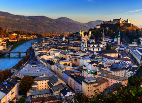 Luftaufnahme einer historischen Stadt am Fluss mit Brücken, Dom- und Kirchtürmen sowie einer Burg auf einem Hügel; dahinter erstrecken sich Berge.