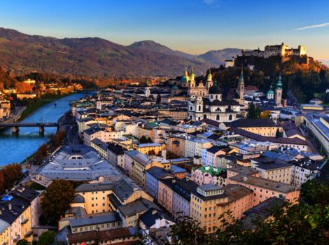 Luftaufnahme einer historischen Stadt am Fluss mit Brücken, Dom- und Kirchtürmen sowie einer Burg auf einem Hügel; dahinter erstrecken sich Berge.