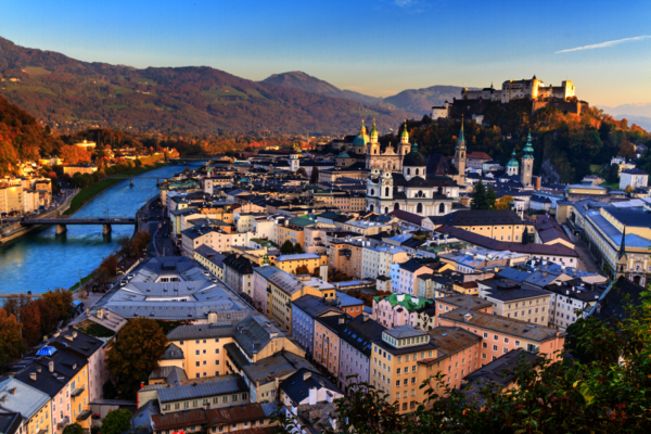 Aerial view of Salzburg, Austria: a river winds through the city, crossing bridges among dense pastel rooftops and church spires; a hilltop fortress overlooks the town, with mountains rising in the distance.