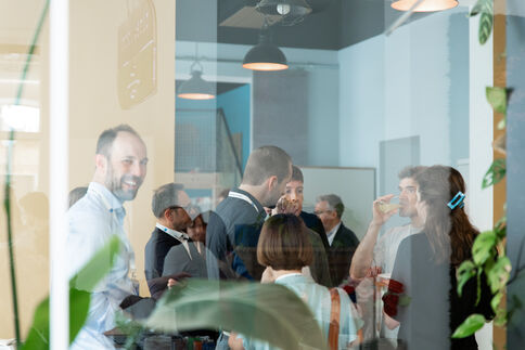 Gruppe von Berufstätigen bei einem Networking-Event in einem modernen Büro-Café. Die Menschen unterhalten sich und halten Getränke; die Szene wirkt durch eine Glaswand mit Pflanzen teilweise sichtbar.