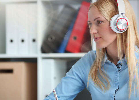 Eine junge Frau mit Kopfhörern sitzt an einem Schreibtisch, arbeitet am Laptop und schreibt mit einem Stift; im Hintergrund stehen Ordnerregale.