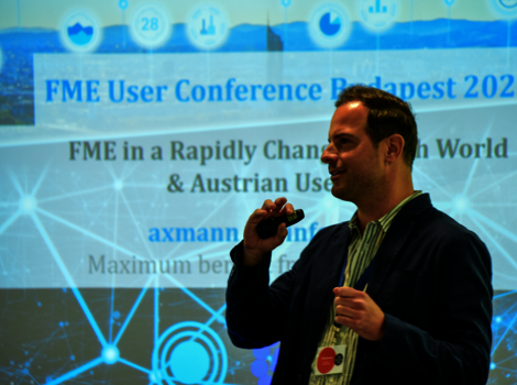 Ein Mann hält ein Mikrofon vor einem Vortrag bei der FME User Conference Budapest 2026. Im Hintergrund eine Projektionswand mit dem Konferenztitel und digitalen Netzwerkgrafiken.