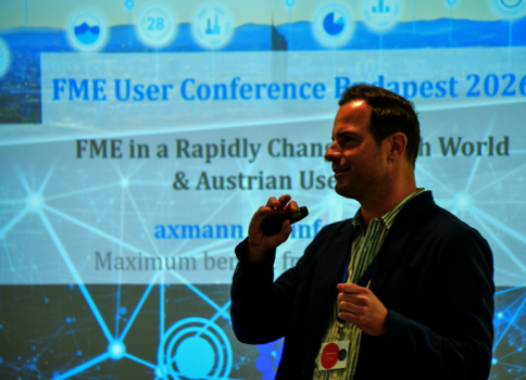 Ein Mann hält ein Mikrofon vor einem Vortrag bei der FME User Conference Budapest 2026. Im Hintergrund eine Projektionswand mit dem Konferenztitel und digitalen Netzwerkgrafiken.