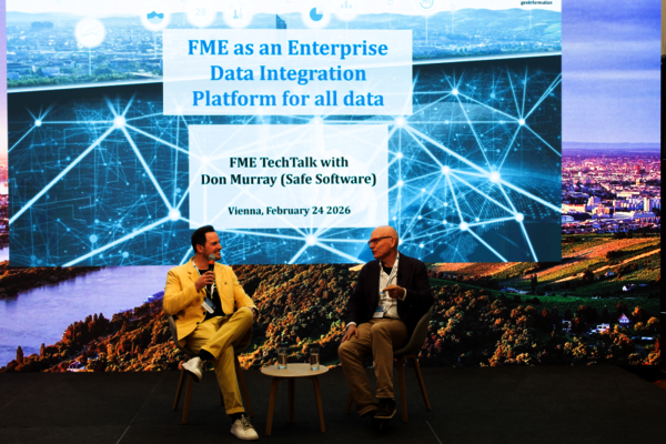 Zwei Männer sitzen auf einer Bühne im Gespräch, jeder hat ein Mikrofon. Hinter ihnen eine große Projektion mit dem Vortragstitel 'FME as an Enterprise Data Integration Platform for all data' und Details zum FME TechTalk in Wien am 24. Februar 2026.