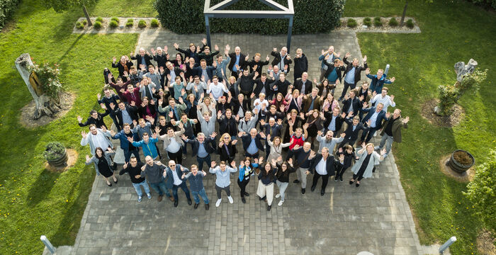 Overhead view of a large, diverse group of people gathered on a gray stone courtyard in a manicured garden, forming a circular cluster and looking up at the camera, with green lawn, trimmed hedges, statues, and a metal framework in the background.