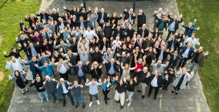 Vogelperspektive auf eine große Gruppe Menschen, die in einem Kreis auf einer gepflasterten Fläche in einem Garten stehen. Alle schauen nach oben und heben die Hände. Umgeben von grünem Rasen, Skulpturen und einer Metallkonstruktion im Hintergrund.