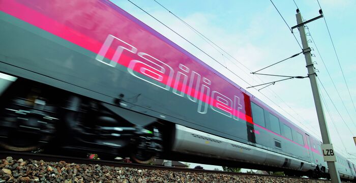 Ein farbenfroher Hochgeschwindigkeitszug mit der Aufschrift Railjet fährt schnell vorbei, rosa und türkis lackiert, auf Schottergleisen; darüber Oberleitungen und ein Mast im Hintergrund.