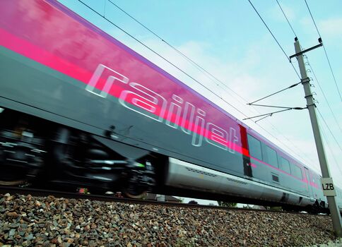 A modern pink-and-teal high-speed passenger train labeled "Railjet" speeds along a gravel railway, under overhead electric lines with a tall power pole in view, seen from a low side angle.