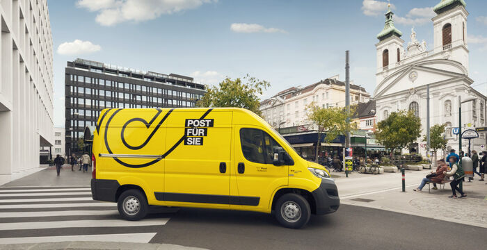 Ein gelber Lieferwagen mit der Aufschrift "POST FÜR SIE" fährt durch eine Stadtkreuzung. Links moderne Bürogebäude, rechts eine weiße Kirche. Fußgänger überqueren den Zebrastreifen, während im Hintergrund weitere Passanten gehen.