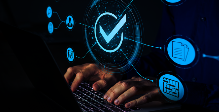 Eine Person tippt auf einem Laptop. Über dem Gerät schwebt ein leuchtendes holographisches Symbol eines blauen Häkchens in einem Kreis, umgeben von digitalen Verbindungen und Symbolen, die Verifikation, Qualitätskontrolle und Datensicherheit andeuten.