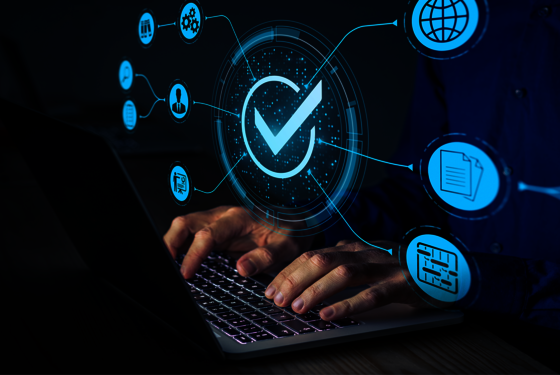 Eine Person tippt auf einem Laptop. Über dem Gerät schwebt ein leuchtendes holographisches Symbol eines blauen Häkchens in einem Kreis, umgeben von digitalen Verbindungen und Symbolen, die Verifikation, Qualitätskontrolle und Datensicherheit andeuten.