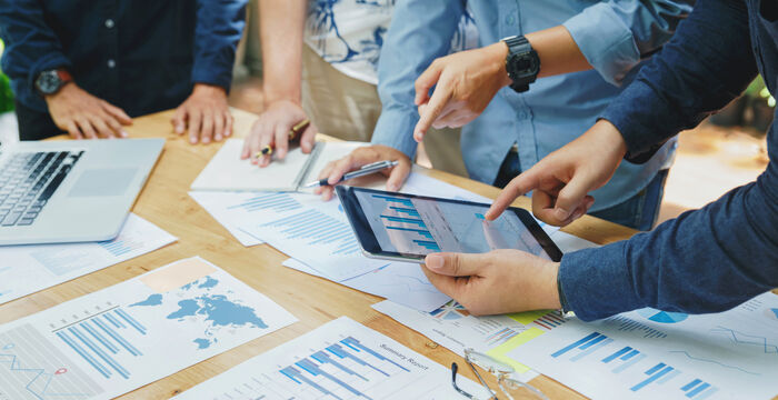 Mehrere Personen arbeiten an einem Tisch mit Unterlagen, Diagrammen, einem Tablet und einem Laptop. Sie analysieren Grafiken und diskutieren Daten.