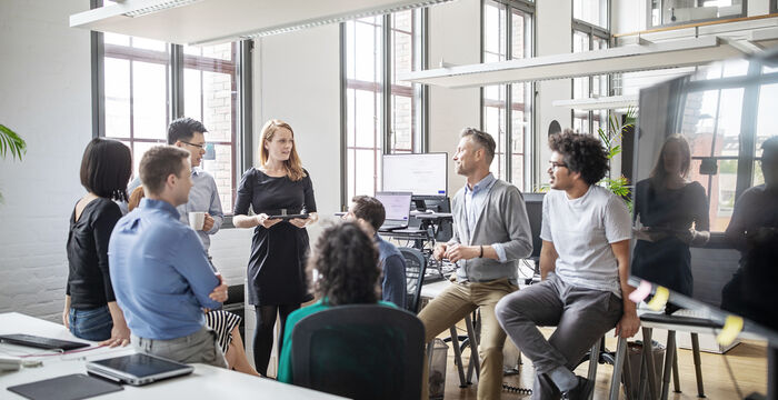 Eine Gruppe von Kolleginnen und Kollegen versammelt sich in einem hellen offenen Büro. Eine Frau hält vorne eine Präsentation, während Mitarbeitende an Tischen arbeiten oder stehen. Große Fenster, moderne Ausstattung, lockere Arbeitsatmosphäre.