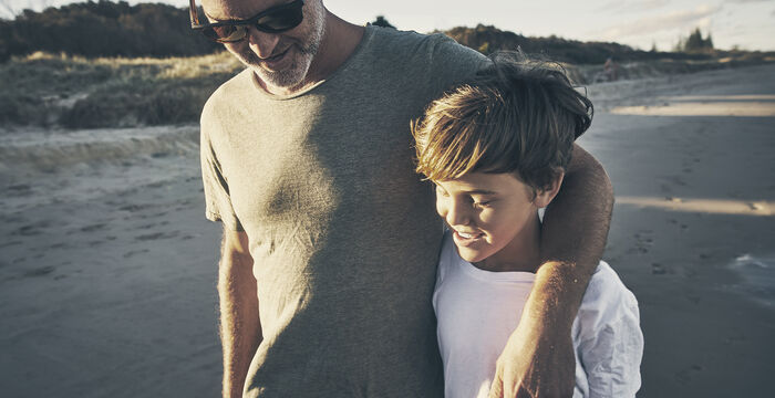 Ein erwachsener Mann mit Sonnenbrille und grauem T‑Shirt geht am Strand mit einem jungen Jungen, der ein weißes Oberteil trägt. Der Mann legt den Arm um dessen Schulter; sie gehen entspannt am Sandstrand entlang.