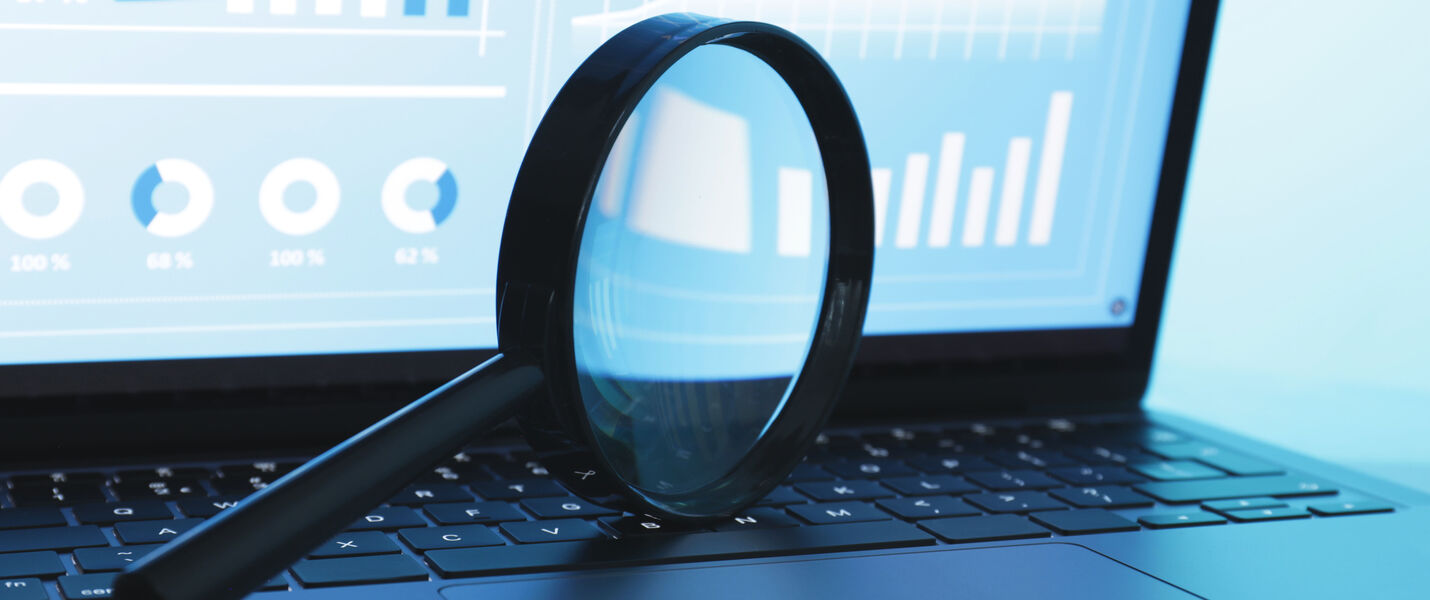 A laptop displaying charts and graphs on the screen, with a magnifying glass lying across the keyboard.