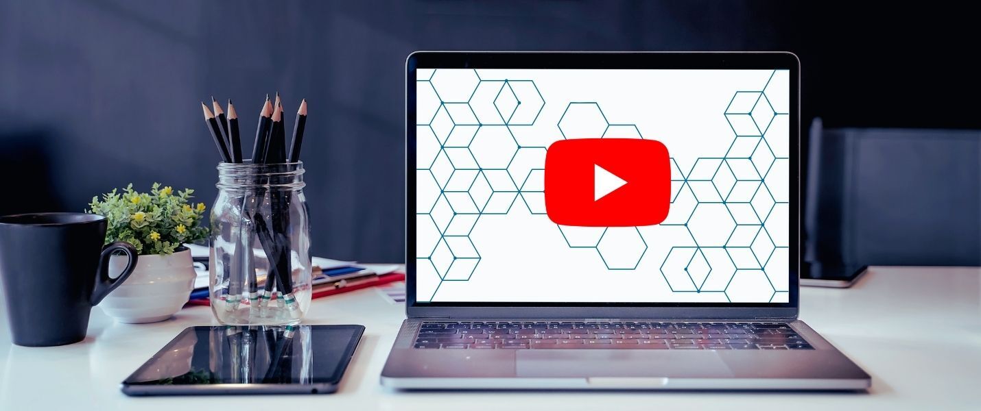 Ein Laptop auf einem Schreibtisch zeigt auf dem Bildschirm das YouTube-Wiedergabe-Symbol; daneben Tablet, Stifte im Glas, Kaffeetasse und Pflanze.