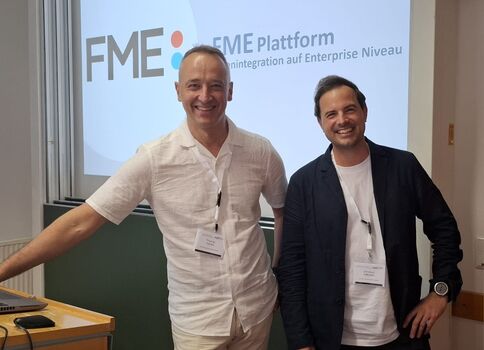 Zwei lächelnde Männer stehen in einem Schulungsraum vor einer Projektionswand mit dem FME-Logo und der Aufschrift „FME Plattform“. Sie tragen Namensschilder; einer in weißem Hemd und beiger Hose, der andere in dunkler Jacke und weißem T-Shirt.