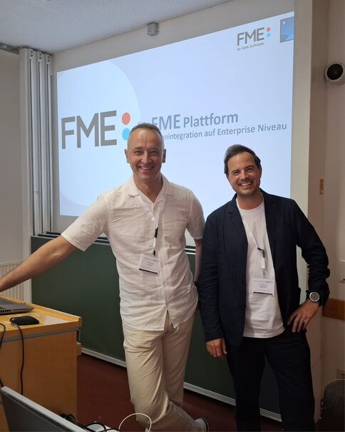 Zwei lächelnde Männer stehen in einem Schulungsraum vor einer Projektionswand mit dem FME-Logo und der Aufschrift „FME Plattform“. Sie tragen Namensschilder; einer in weißem Hemd und beiger Hose, der andere in dunkler Jacke und weißem T-Shirt.