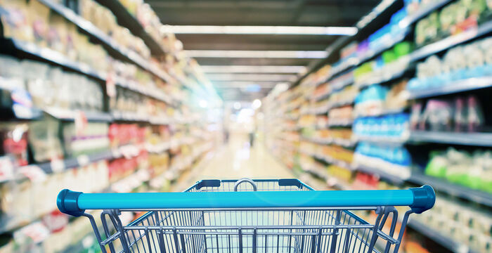 Perspektive hinter einem Einkaufswagen in einem hell beleuchteten Supermarkt. Links und rechts ziehen Regale mit bunten Produkten vorbei, während der lange Gang ins Zentrum des Ladens führt.