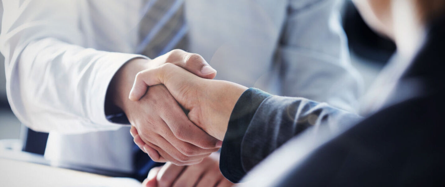 Two business professionals shake hands across a desk in an office, signaling an agreement or partnership.