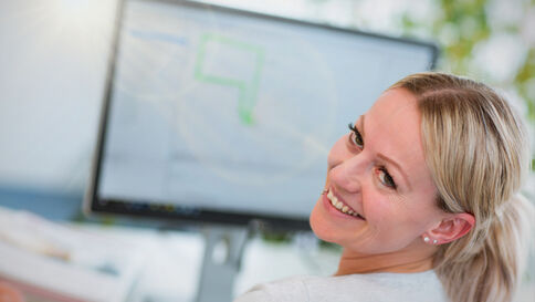 Eine lächelnde Frau sitzt am Arbeitsplatz; im Hintergrund sieht man einen Computerbildschirm mit einer Grafik.