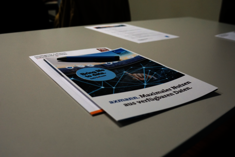 Auf einem Tisch liegt eine Broschüre. Das Titelblatt zeigt eine blaue Netzgrafik und den Satz „Maximalen Nutzen aus verfügbaren Daten“, der auf datenbasierte Nutzung hinweist.