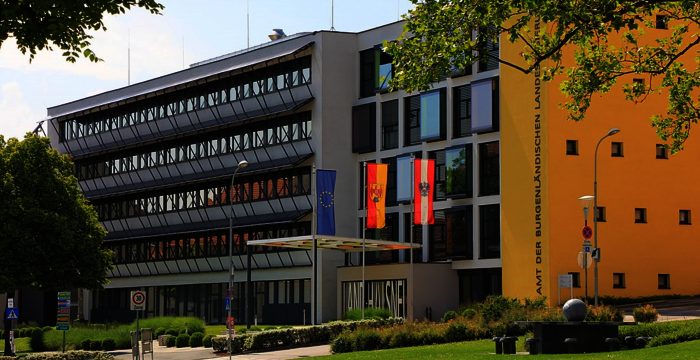 Modernes Verwaltungsgebäude mit Glasfassade und mehreren Flaggen (EU, Deutschland und weitere) vor dem Eingang. Im Vordergrund gepflegte Grünfläche, Bäume spenden Schatten; rechts eine Beschilderung der Behörde.