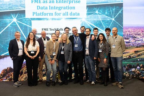 Eine Gruppe von ca. 15 Personen steht nebeneinander auf einer Bühne bei einer Technologie-Konferenz und blickt in die Kamera. Sie tragen Namensschilder; im Hintergrund ist eine große Leinwand mit dem Text 'FME as an Enterprise Data Integration Platform for all data' zu sehen.