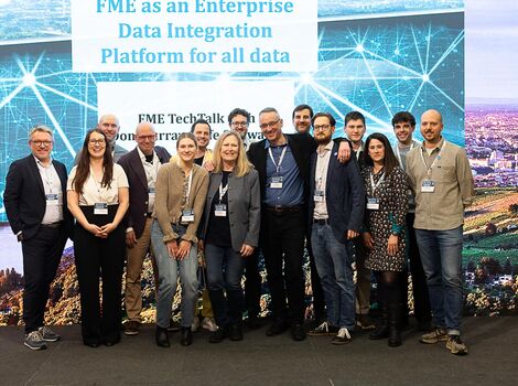 Eine Gruppe von ca. 15 Personen steht nebeneinander auf einer Bühne bei einer Technologie-Konferenz und blickt in die Kamera. Sie tragen Namensschilder; im Hintergrund ist eine große Leinwand mit dem Text 'FME as an Enterprise Data Integration Platform for all data' zu sehen.