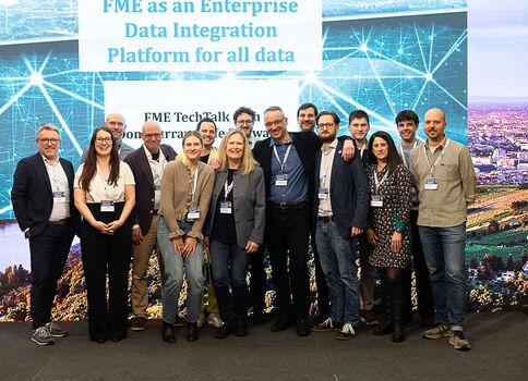 Eine Gruppe von ca. 15 Personen steht nebeneinander auf einer Bühne bei einer Technologie-Konferenz und blickt in die Kamera. Sie tragen Namensschilder; im Hintergrund ist eine große Leinwand mit dem Text 'FME as an Enterprise Data Integration Platform for all data' zu sehen.