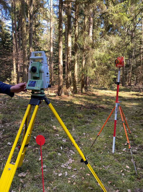 Vermessungsgeräte im Wald: Im Vordergrund ein Totalstation auf einem gelben Dreibein-Stativ. Links daneben ein rotes Zielschild auf einem Stab. Im Hintergrund steht ein zweites Stativ mit einem Messgerät auf einem rot-weiß gestreiften Mast; viele Bäume und Laub umgeben die Szene.