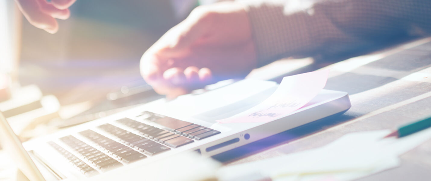A sunlit desk scene featuring an open laptop, scattered papers, pencils, and a ruler, with a person’s hands resting near the keyboard, suggesting active work or study.