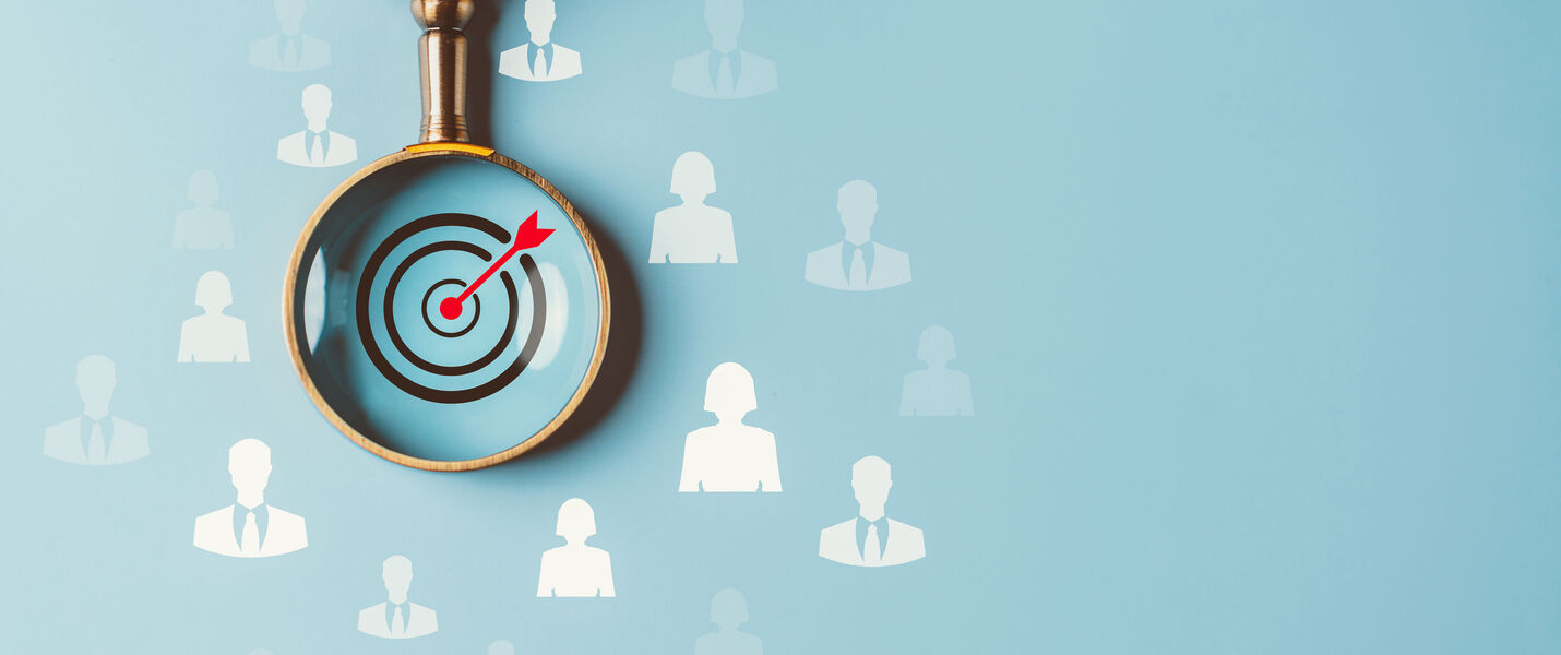 A hand holds a magnifying glass over a blue background, revealing a bullseye target with a red arrow hitting its center, symbolizing targeting a specific audience.