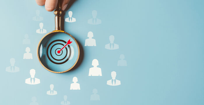 Eine Hand hält eine goldene Lupe, durch die eine Zielscheibe mit rotem Pfeil im Zentrum sichtbar ist; im Hintergrund schweben weiße Personen-Symbole – Symbol für Zielgruppenauswahl oder gezielte Ansprache.