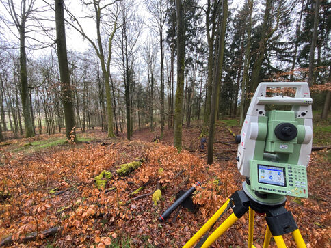Vermessungsgerät (Totalstation) auf Stativ im Laubwald; braunes Herbstlaub bedeckt den Boden, im Hintergrund stehen Bäume.
