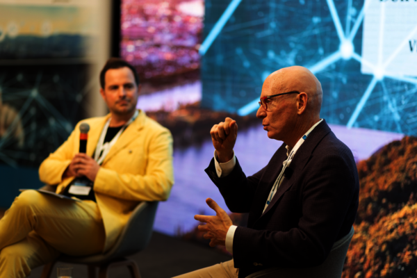 Zwei Männer sitzen auf einer Bühne in einer Diskussionsrunde. Links hält ein Mann in gelber Jacke ein Mikrofon, rechts ein älterer Mann mit Brille in dunklem Blazer macht eine erklärende Handgeste vor einem Hintergrund mit Bildschirmen und Mustern.