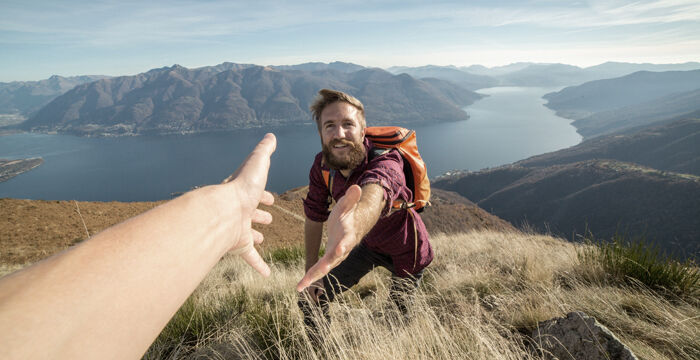 Ein Wanderer mit Rucksack reicht dem Fotografen die Hand, während sie auf einem Bergkamm mit Blick auf einen See und umliegende Bergketten im Hintergrund stehen.