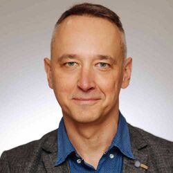 Porträt eines Mannes mittleren Alters mit kurzen grauen Haaren, blauem Hemd und grauem Blazer. Er schaut in die Kamera und lächelt leicht vor hellem Hintergrund.