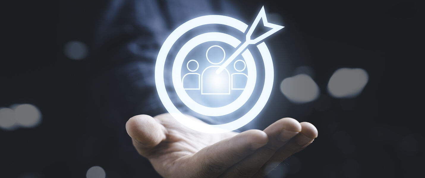A glowing white holographic icon shows a target with an arrow hitting the center, surrounding silhouettes of people, hovering above an open hand—symbolizing targeting or focusing on an audience.
