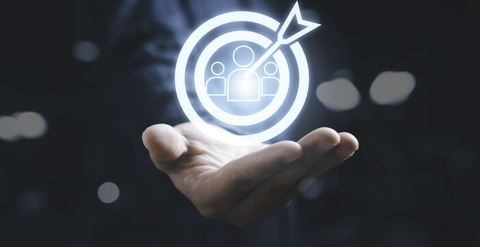 A glowing white holographic icon shows a target with an arrow hitting the center, surrounding silhouettes of people, hovering above an open hand—symbolizing targeting or focusing on an audience.