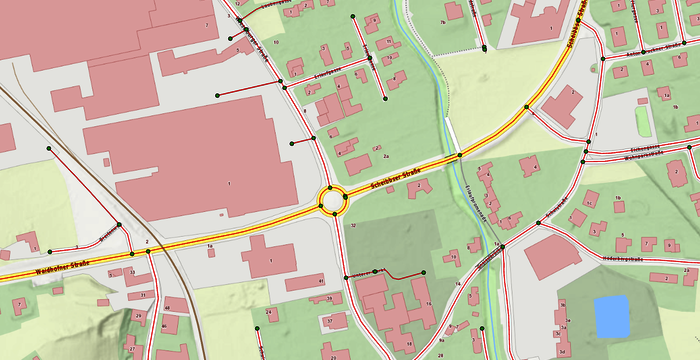 Stadtplan mit zentralem Kreisverkehr an der Kreuzung mehrerer Straßen. Umgeben sind rote Gebäudeblöcke, links größere Blockstrukturen, rechts und unten kleinere Häuser. Grünflächen und Parks dominieren das Gebiet; unten rechts befindet sich ein kleiner blauer Teich, ein Wasserlauf zieht sich durch die Mitte.
