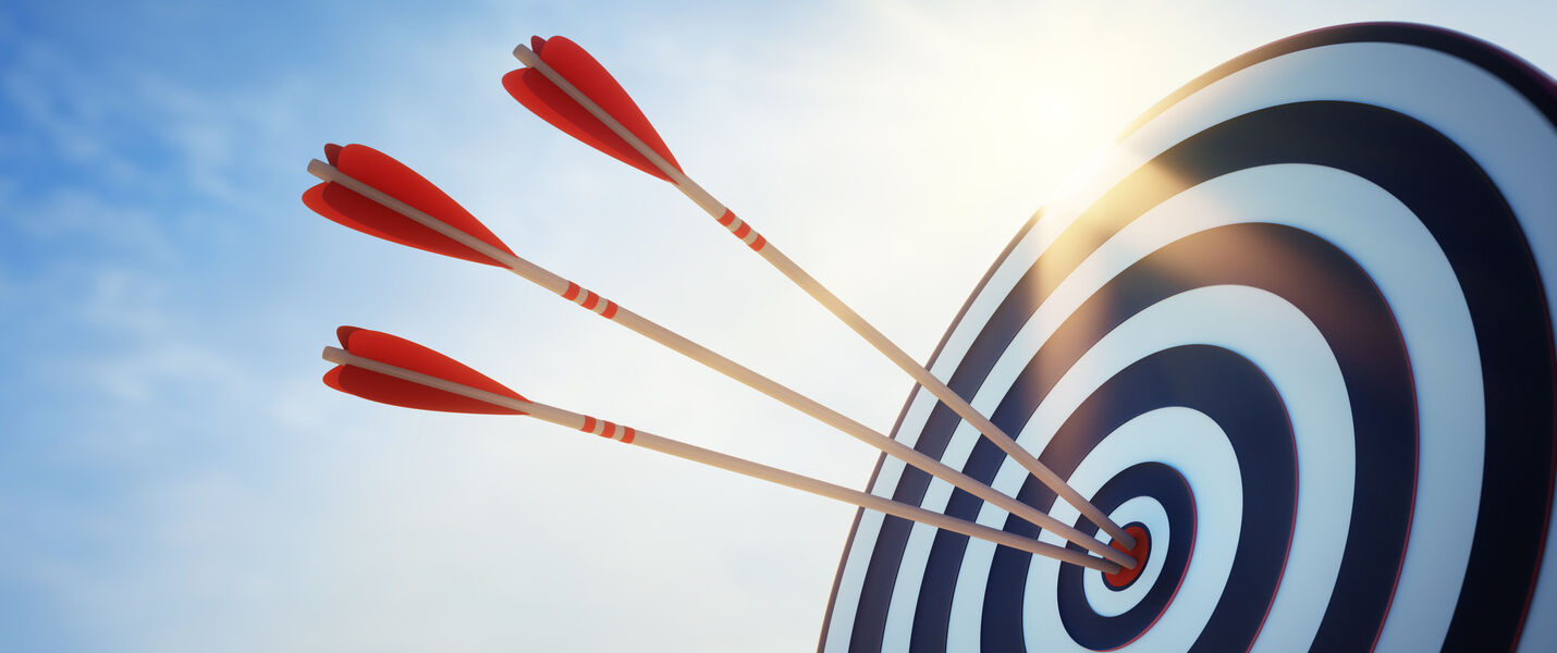 Three red darts embedded in the bullseye of a blue-and-white target, with a bright sky and sun glare behind.