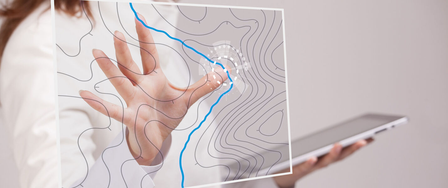 Eine Person hält ein Tablet und berührt eine transparente Oberfläche mit topografischen Konturen; eine blaue Linie markiert einen Weg durch die Konturen.